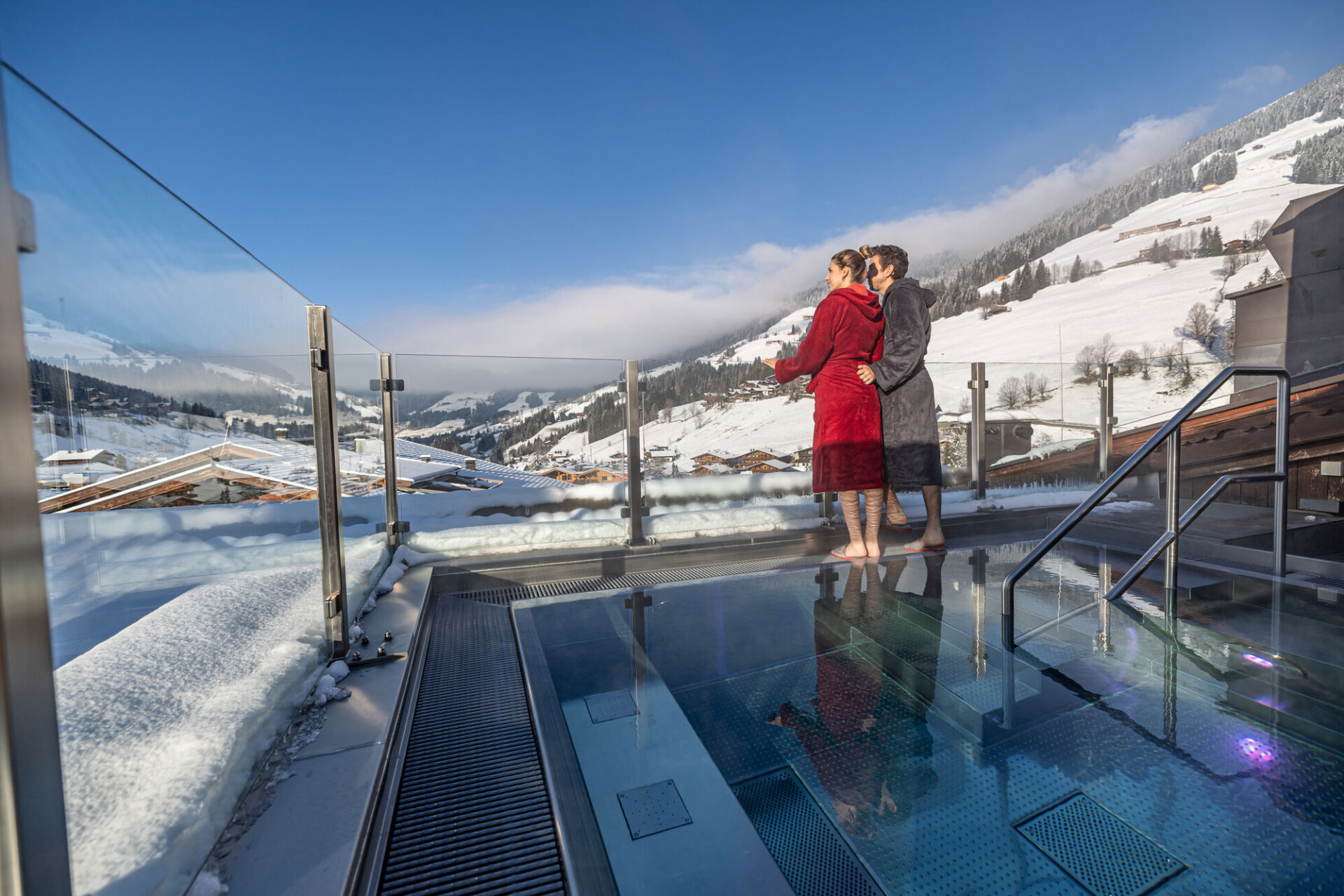 Zwei Menschen in Bademänteln stehen an einer Glasbarriere auf dem Dach des Galtenberg Resorts und blicken auf eine schneebedeckte Berglandschaft neben einem schimmernden Pool.