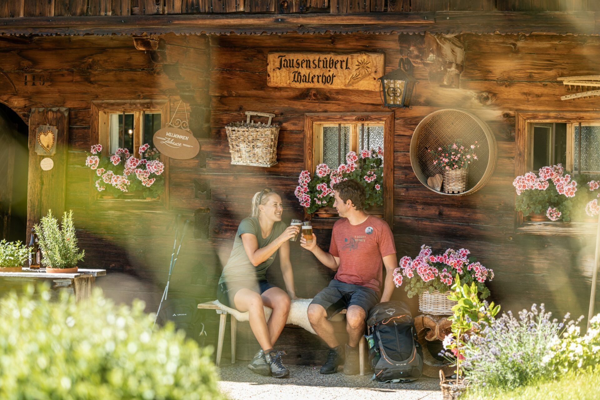 Zwei Personen sitzen vor einer Blockhütte und stoßen mit Bier an.