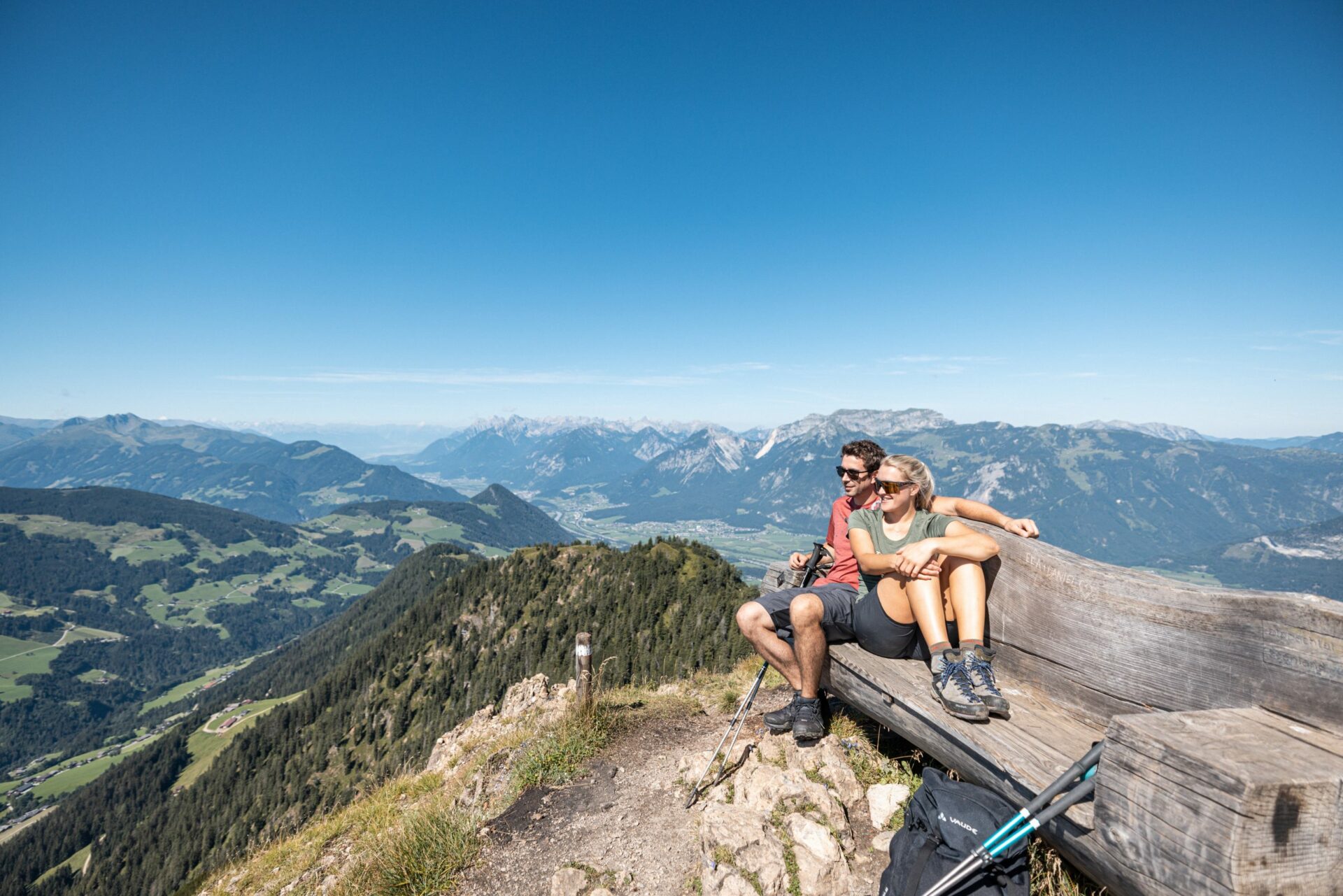 Paar sitzt auf einer Bank auf einem Gipfel und schaut auf die Berge.
